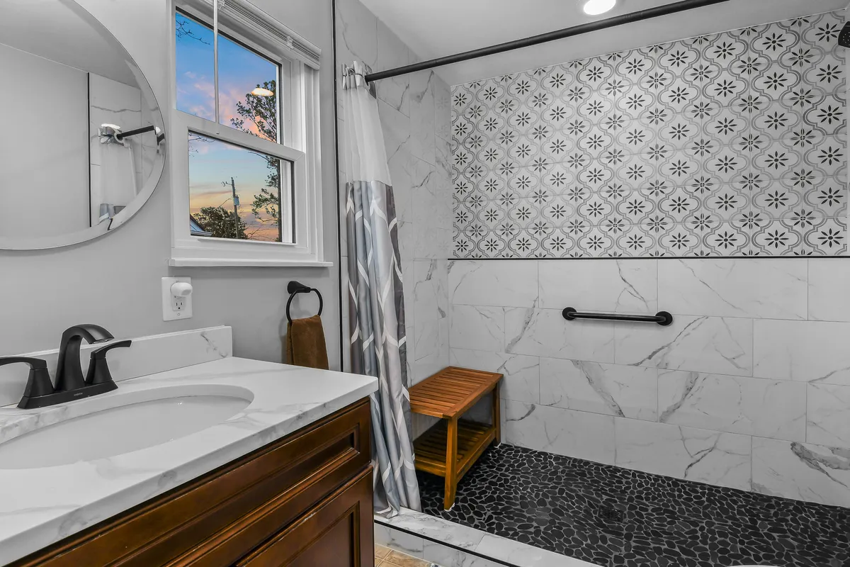 A modern, tranquil bathroom scene featuring a marble vanity, decorative shower wall, wooden shower bench, and a round mirror. The overall look evokes a spa-like experience, as described in the master suite feature.