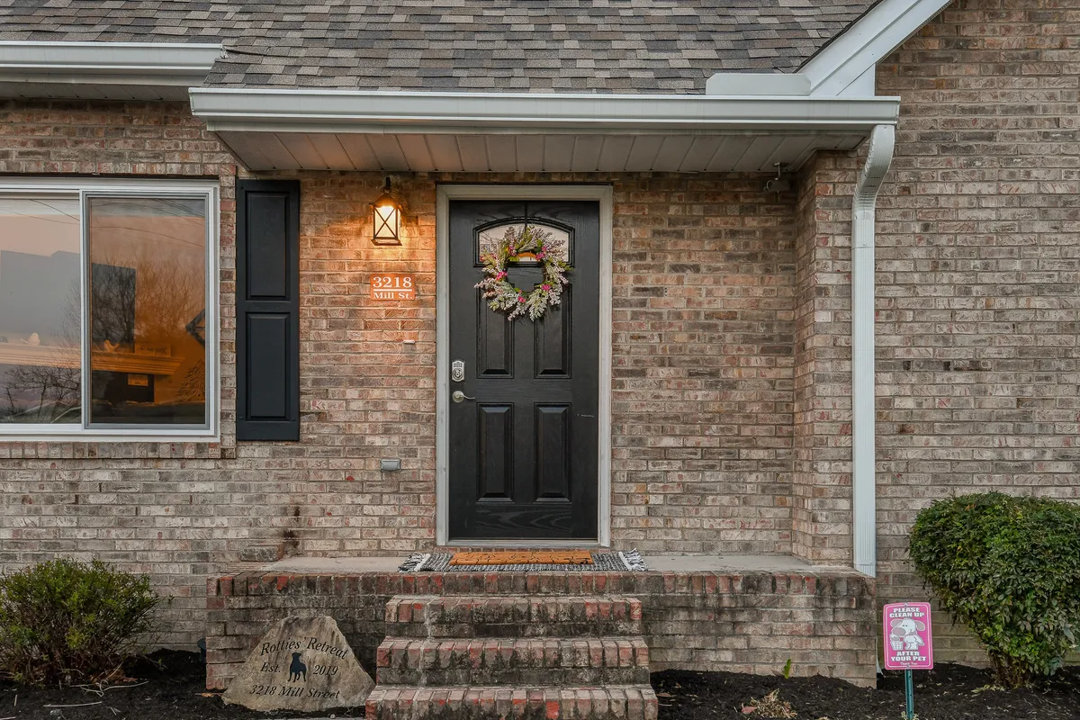 The front door of the house, decorated with a cheerful wreath, brick facade, reflective window, and landscaped bushes, inviting guests to step inside for family adventures.