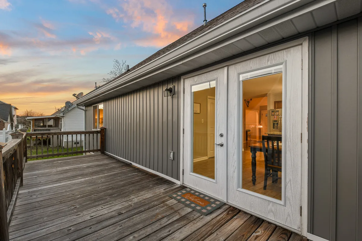 The image captures a wooden deck with double doors leading inside, bathed in the warm glow of a sunset sky. The outdoor space suggests intimate gatherings, relaxation, and the possibility of alfresco meals or drinks under the stars.