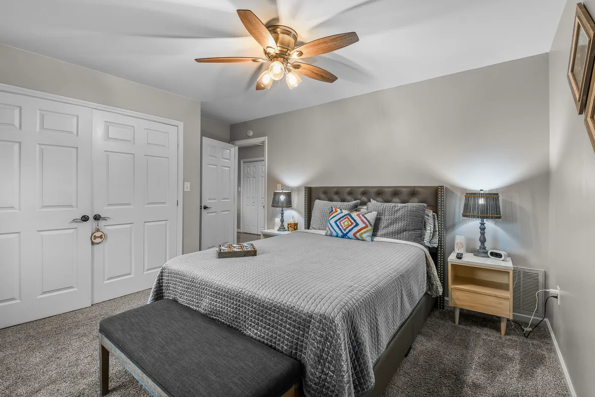 This image showcases a serene bedroom with a comfortable bed, bench, nightstands, ceiling fan, and decorative pillow. The room is bathed in natural light, creating an inviting and restful atmosphere, ideal for relaxation and unwinding.