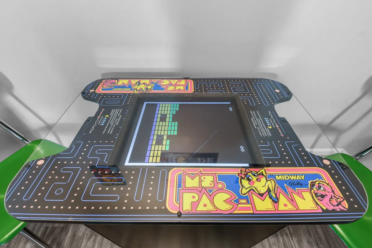 This playful corner features a classic Ms. Pac-Man arcade game, a sleek glass table with green chairs, and vibrant décor. The setup is designed for friendly competition and fun, inviting guests of all ages to engage in gaming sessions and lively social interactions.