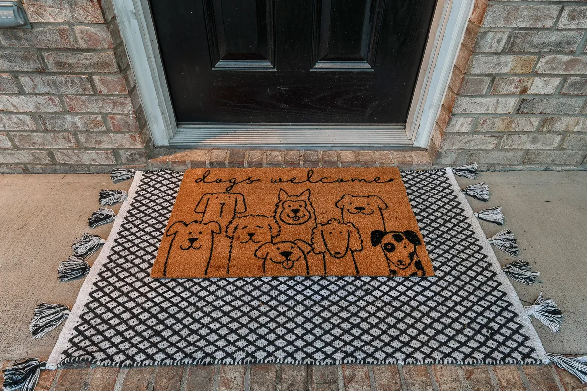 An exterior scene featuring a doormat with playful dog illustrations outside a black front door and a brick wall. The scene radiates a friendly welcome, with the doormat signaling pet-friendliness and inclusivity for guests with dogs.