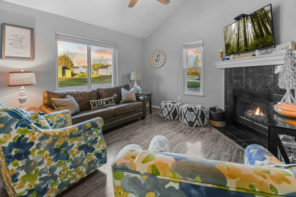 This inviting living room scene features a comfortable brown leather sofa, colorful floral chairs, a tranquil gas log fireplace with wood accents, large windows letting in natural light, and a TV displaying a peaceful forest scene. The arrangement encourages lounging, conversation, and cozy movie nights.
